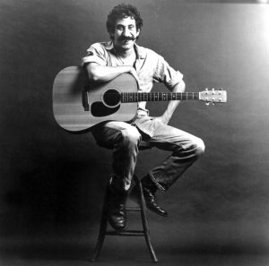 A black and white photo of a mustached man sitting on a stool and resting his arm on a guitar