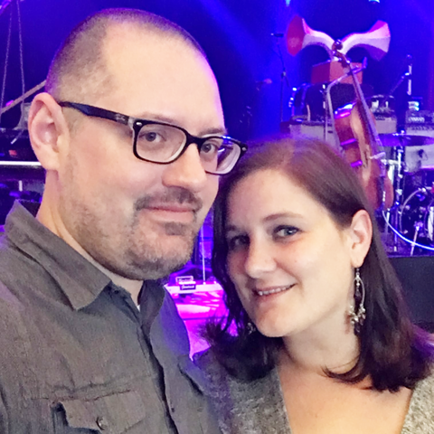 Woman with medium brown hair and man with glasses standing in front of a concert stage