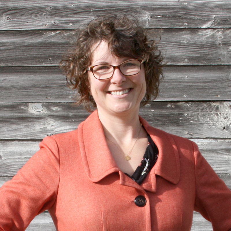 A woman, Nancy Brooks, in a salmon-colored jacket is smiling in front of a wooden wall
