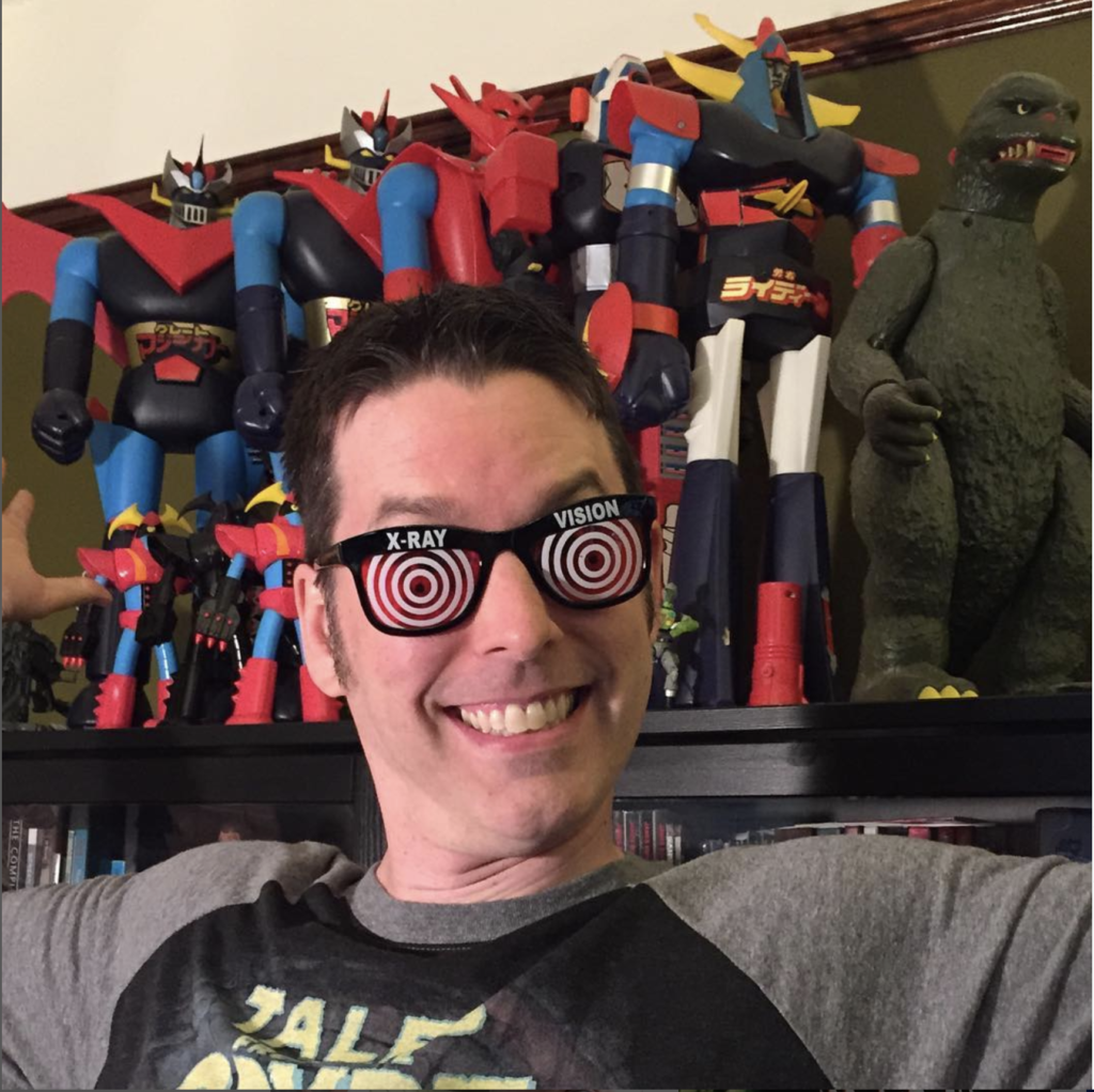 Grinning man wearing toy glasses sits in front of wall of robot toys.