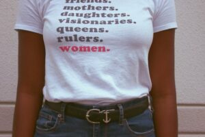 A person wears a white shirt that says: Friends. Mothers. Daughters. Visionaries. Queens. Rulers. Women. Women is in red text and has a tiny heart next to it instead of a period.